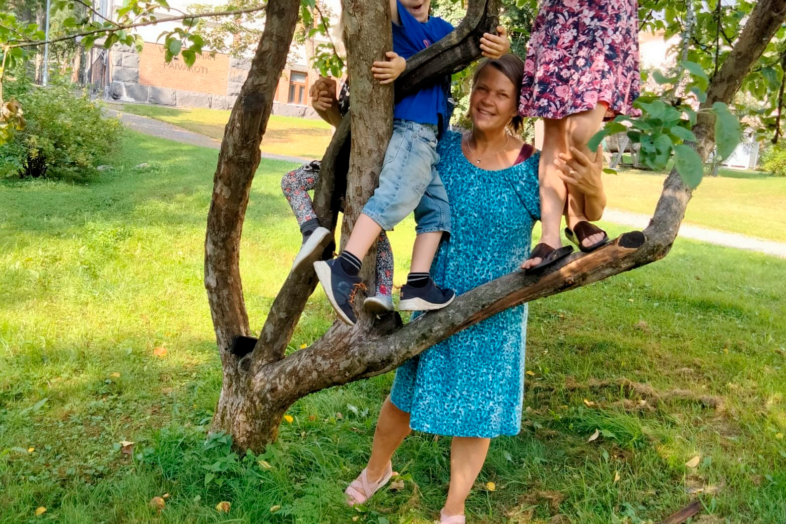 Kuntoutussuunnittelija Merja Smahl hymyilee puunjuurella sinisessä mekossa. Hänen kolme lastenlastaan kiipeilee puunoksilla ja Merja pitää syleillen kiinni lasten jaloista.