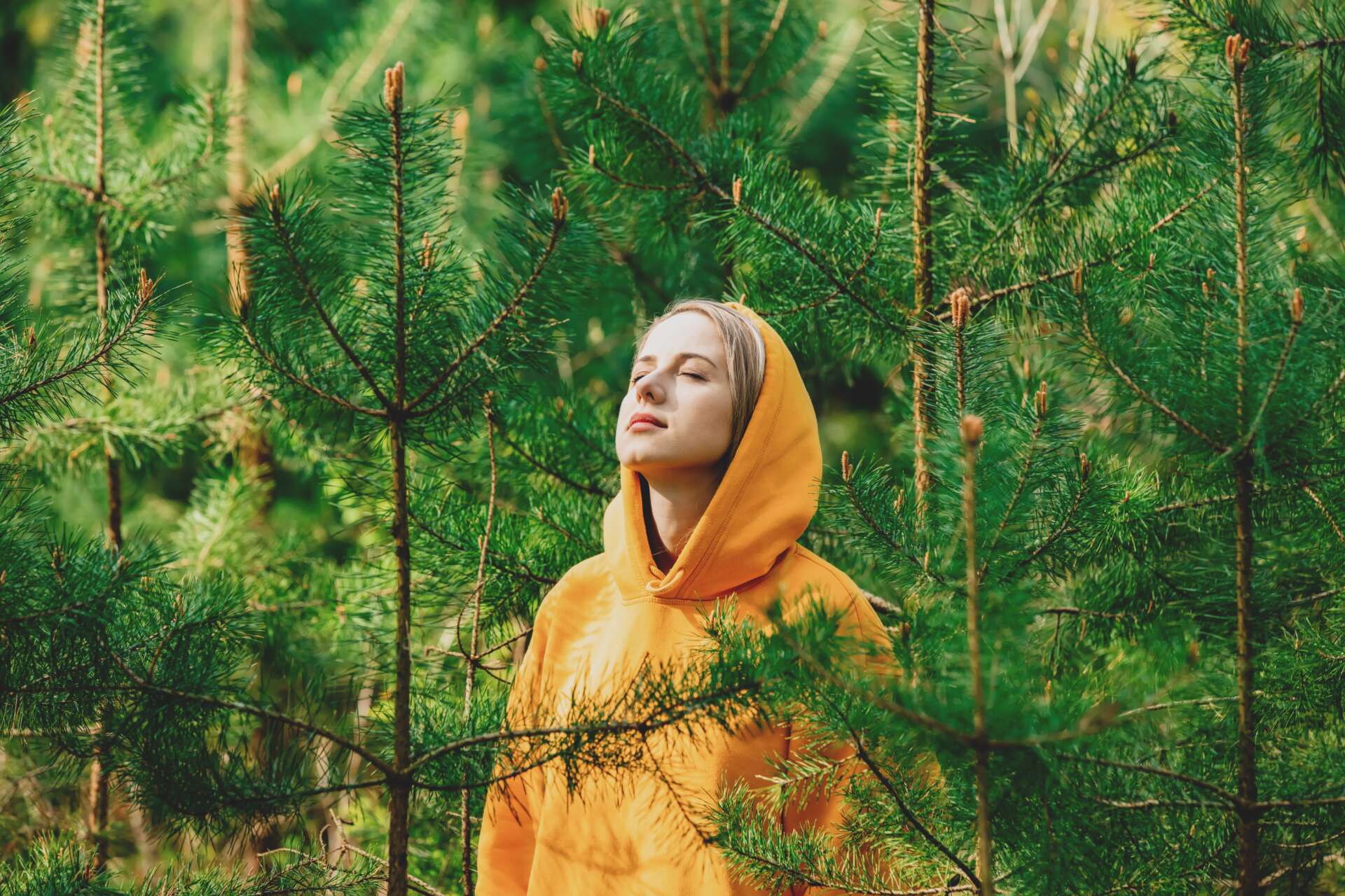 Keltaiseen huppariin pukeutunut tyttö seisoo silmät kiinni mäntyjen keskellä kuuntelemassa luonnon ääniä.