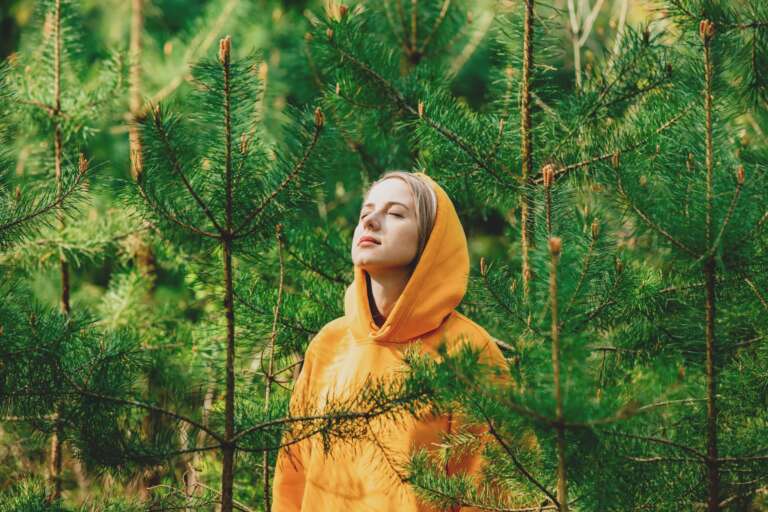 Keltaiseen huppariin pukeutunut tyttö seisoo silmät kiinni mäntyjen keskellä kuuntelemassa luonnon ääniä.