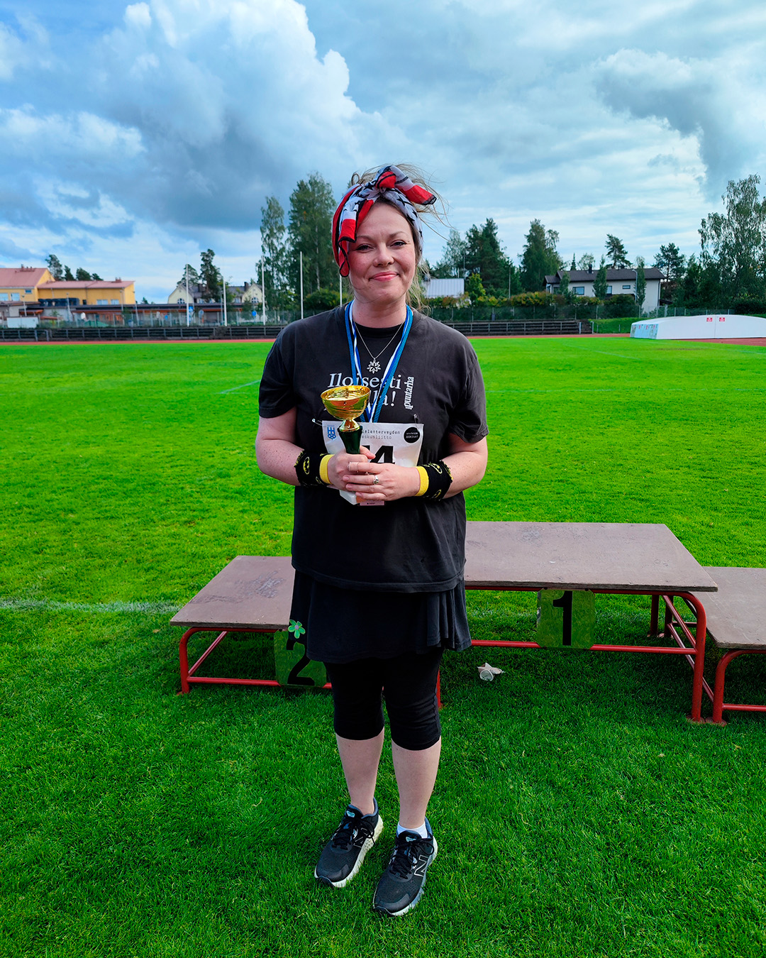 Saara Kananen seisoo urheilukentällä palkintopallin edessä pokaali kädessään ja hymyilee. Hänellä on hiukset päälaella nutturalla, huivi rusettina, mustat trikoot ja musta lyhythihainen paita.