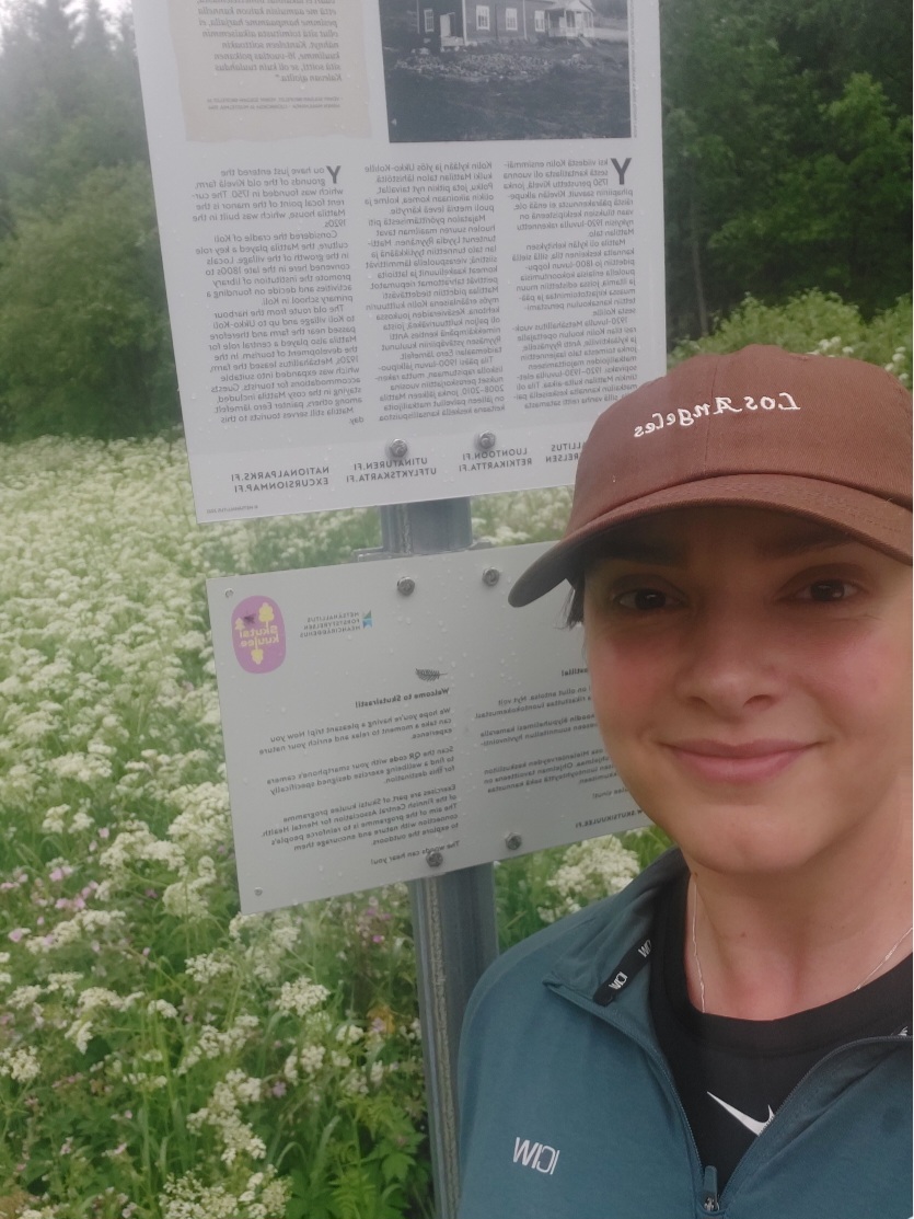 Julia Sillanpää hymyilee Kolin kansallispuiston Skutsirasti-taulun edessä takanaan kukkaketo.