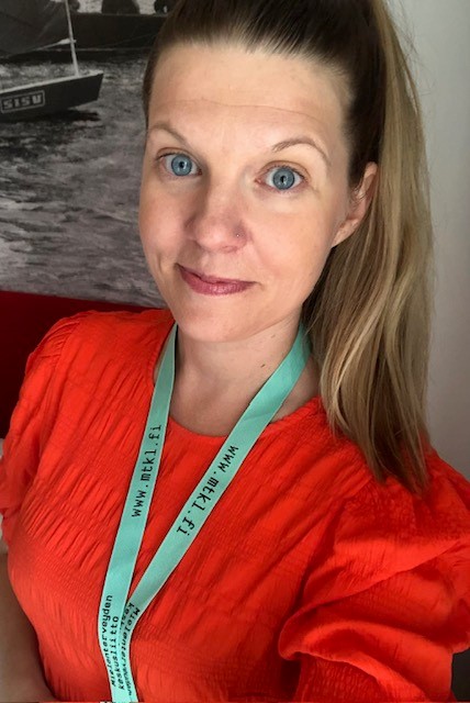 Riikka Mettälän selfie. Hänellä on punainen paita, Mielenterveyden keskusliiton vihreä avainnauha kaulassaan ja tummat pitkät hiukset ponnarilla. Riikka hymyilee.