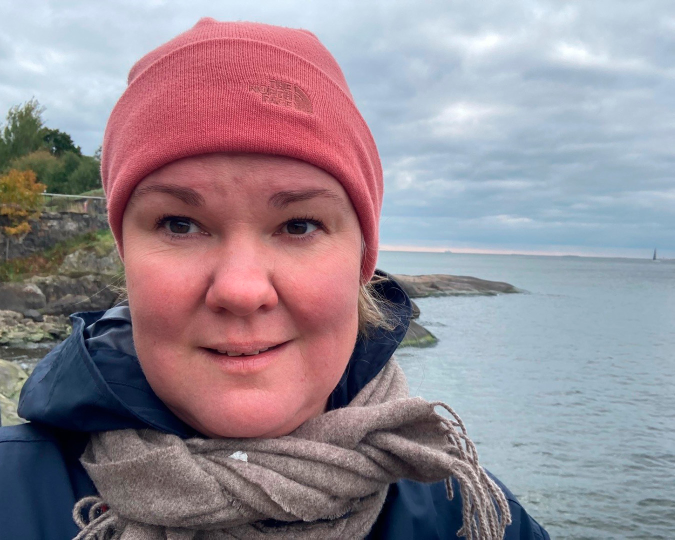 Selfiekuvassa Paula Paloheimo seisoo merenrannalla ja hymyilee. Hänellä on vaaleanpunainen pipo, ruskea kaulaliina ja sininen takki. 