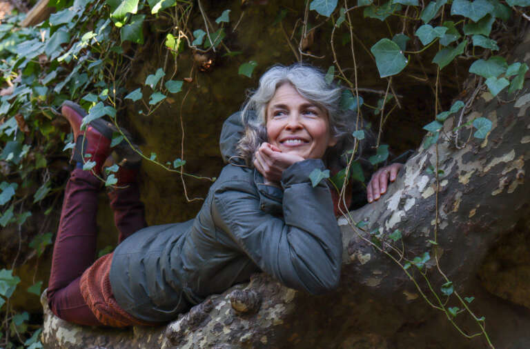Irina Björklund makaa puunoksalla ja nojaa leukaa käteensä. Hänen takanaan köynnökset laskeutuvat kallion seinämältä.