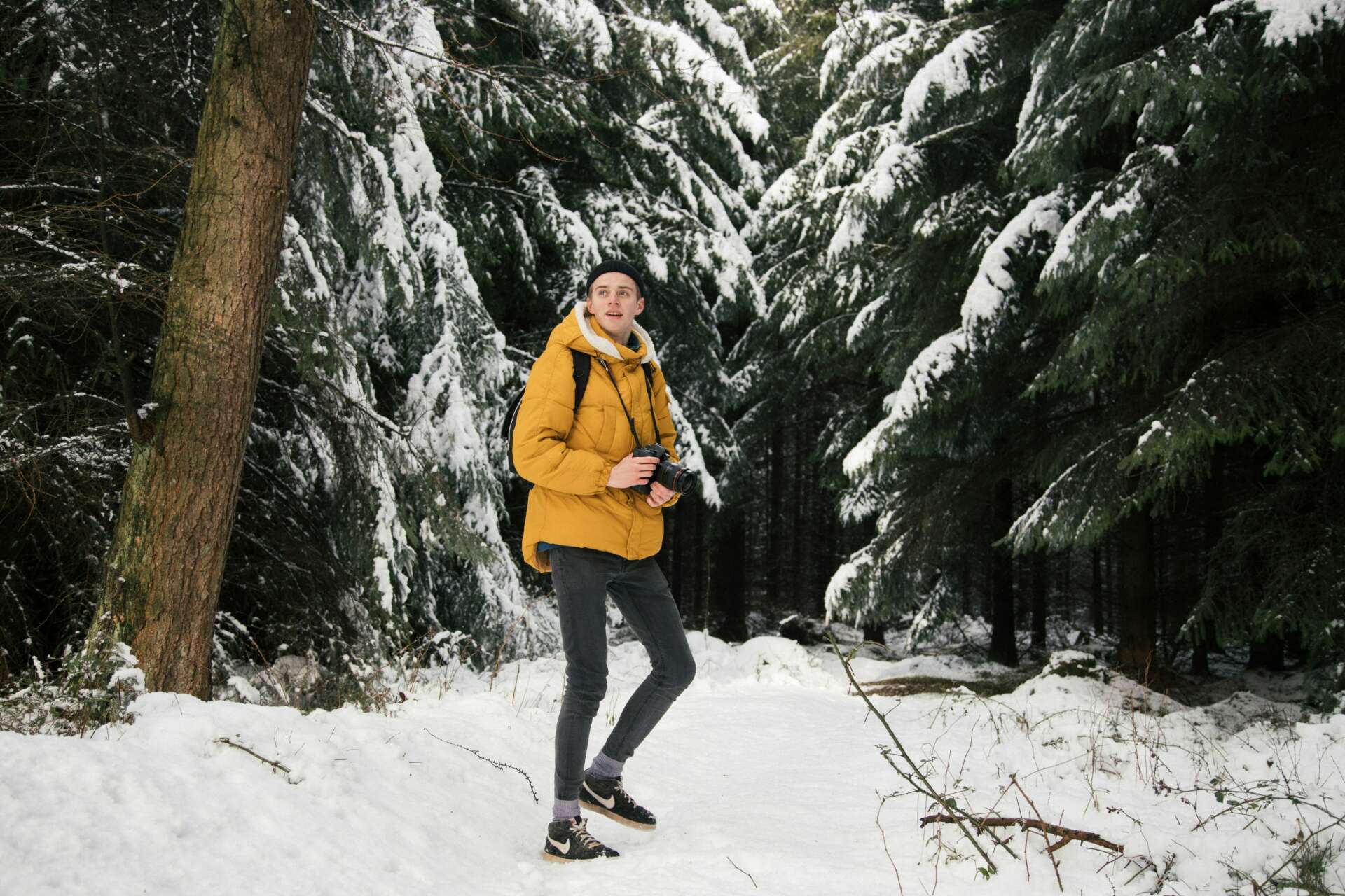 Keltaiseen takkiin pukeutunut nuori mies kävelee lumisten puiden välissä. Hänellä on mukanaan kamera.