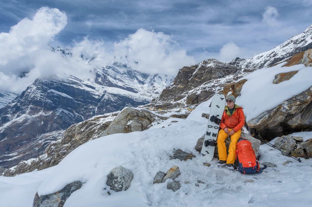 Teemu Suominen istuu kivenlohkareella lumisilla vuorilla vieressään lumilauta ja takana vuoristo, jonka huiput koskettavat pilviä.