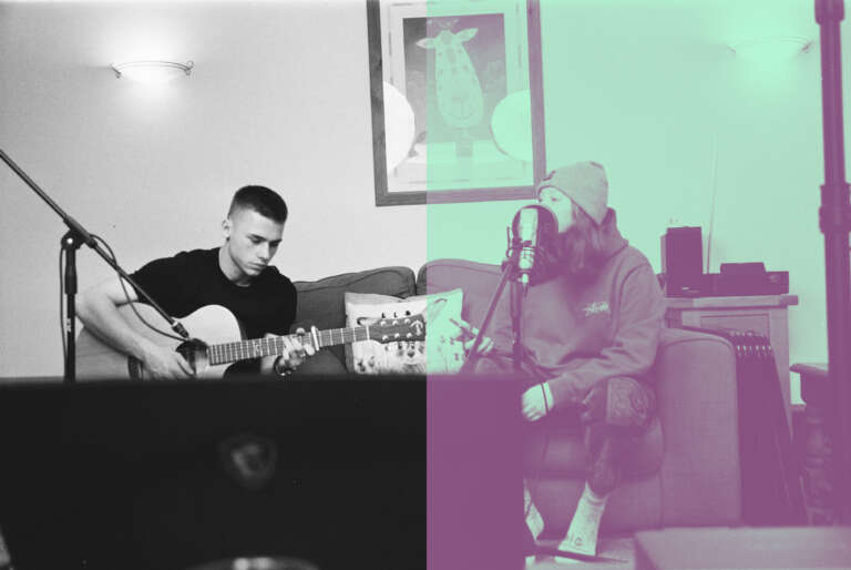Kaksi ihmistä istuu sohvalla. Toinen soittaa kitaraa ja toinen laulaa mikrofoniin.